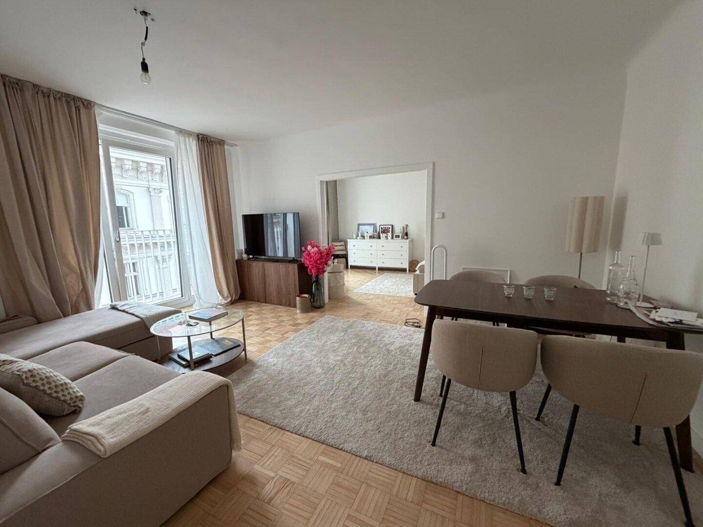Heller Wohn-Essbereich mit Parkettboden, beigem Sofa und Esstisch, sowie Zugang zu einem französischen Balkon.