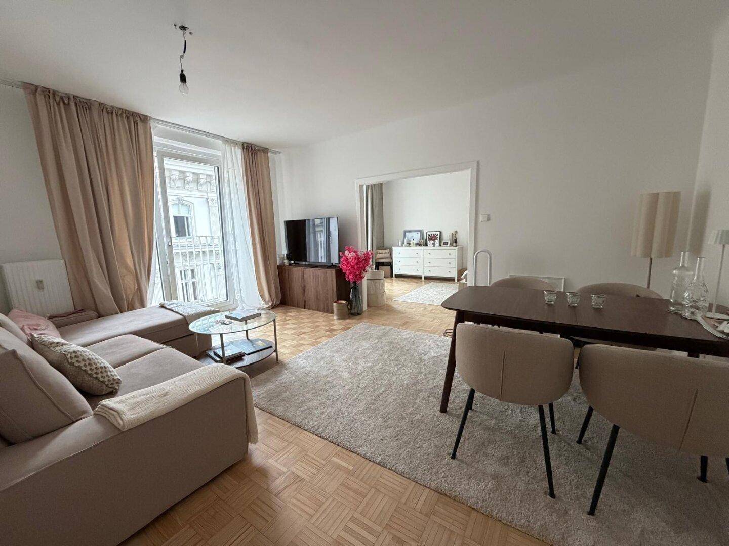 Heller Wohn-Essbereich mit Parkettboden, beigem Sofa und Esstisch, sowie Zugang zu einem französischen Balkon.