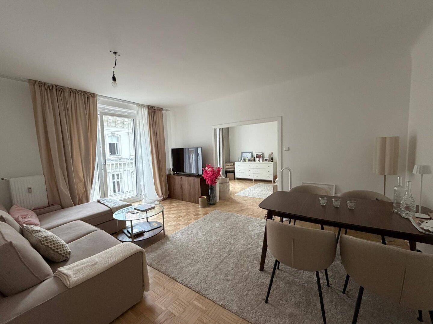 Heller Wohn-Essbereich mit Parkettboden, beigem Sofa und Esstisch, sowie Zugang zu einem französischen Balkon.