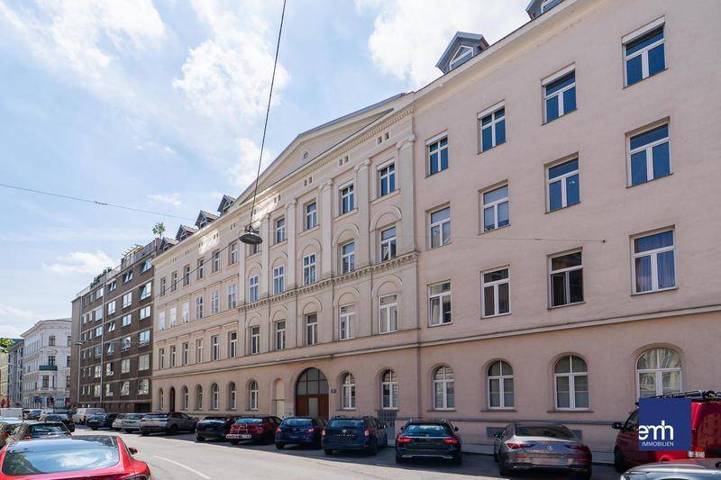 Historische Fassade eines Mehrfamilienhauses an einer belebten Stadtstraße mit parkenden Autos.