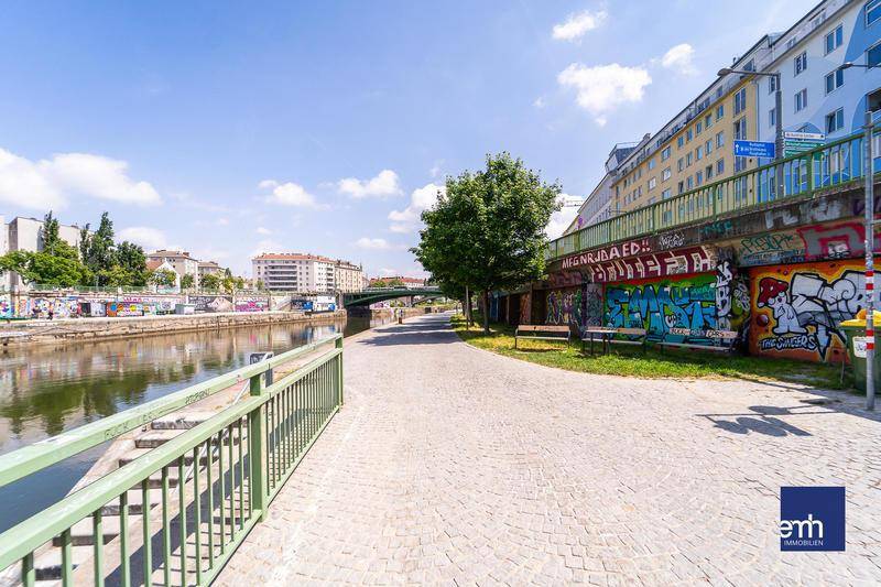 Sonniges Flussufer mit Spazierweg, Graffiti-Wänden und Blick auf die Stadt.