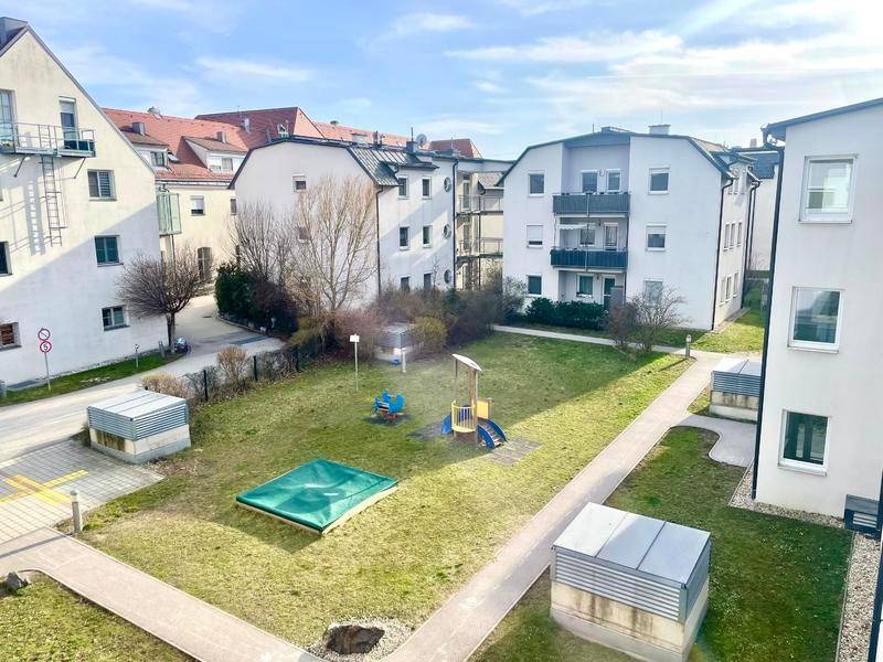 Gepflegter Spielplatz mit Rasenfläche, Sandkasten und Spielgeräten zwischen Wohngebäuden.