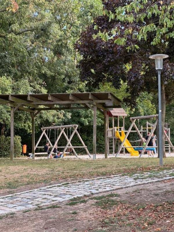 Ein schattiger Spielplatz mit Schaukeln, Rutsche und einer Überdachung, umgeben von Bäumen.