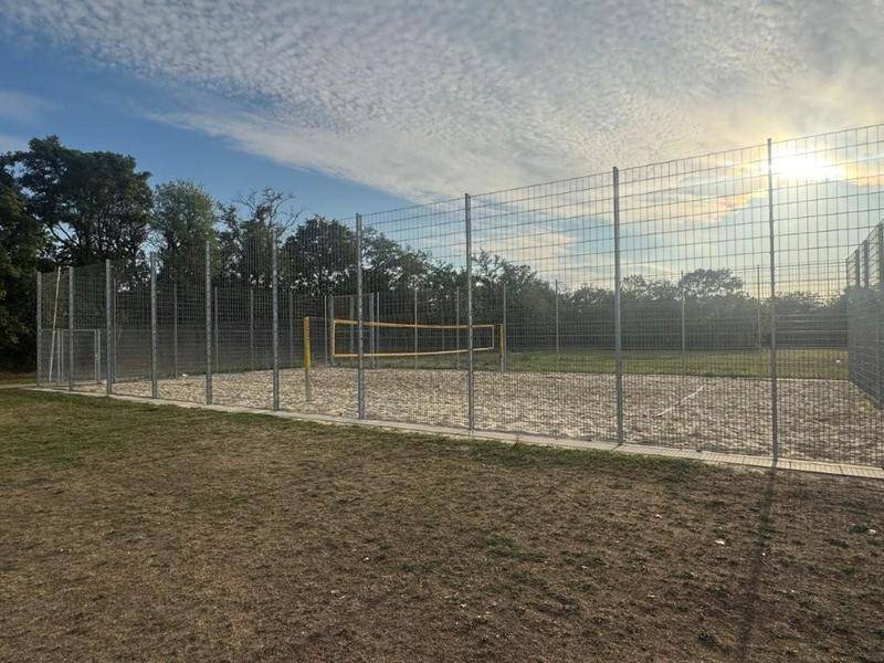 Ein eingezäunter Beachvolleyballplatz mit Sandboden unter blauem Himmel, umgeben von Bäumen.