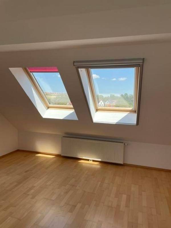 Helles Dachzimmer mit zwei Dachfenstern, eines davon mit rosafarbenem Rollo.