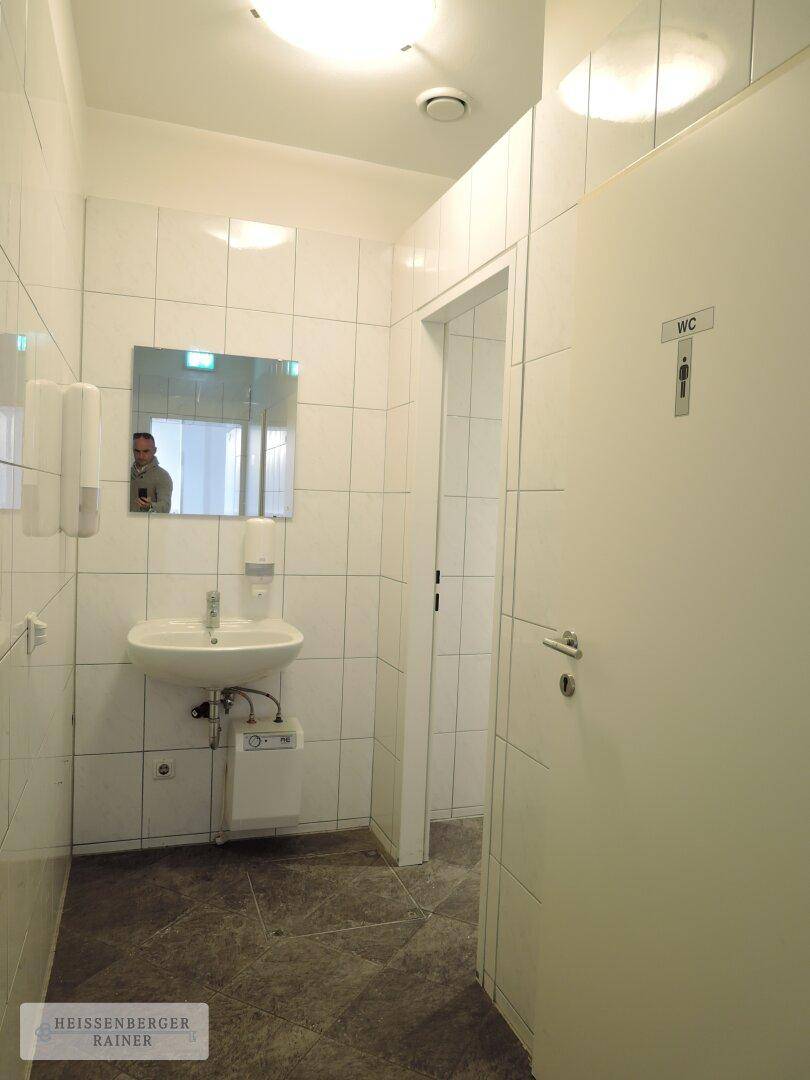 Kleiner, gefliester Toilettenraum mit Waschbecken, Spiegel und WC-Kabinentür.