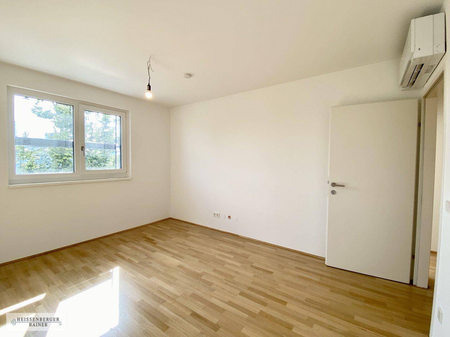 Helles, leeres Zimmer mit Holzboden und großem Fenster, ideal als Schlafzimmer.