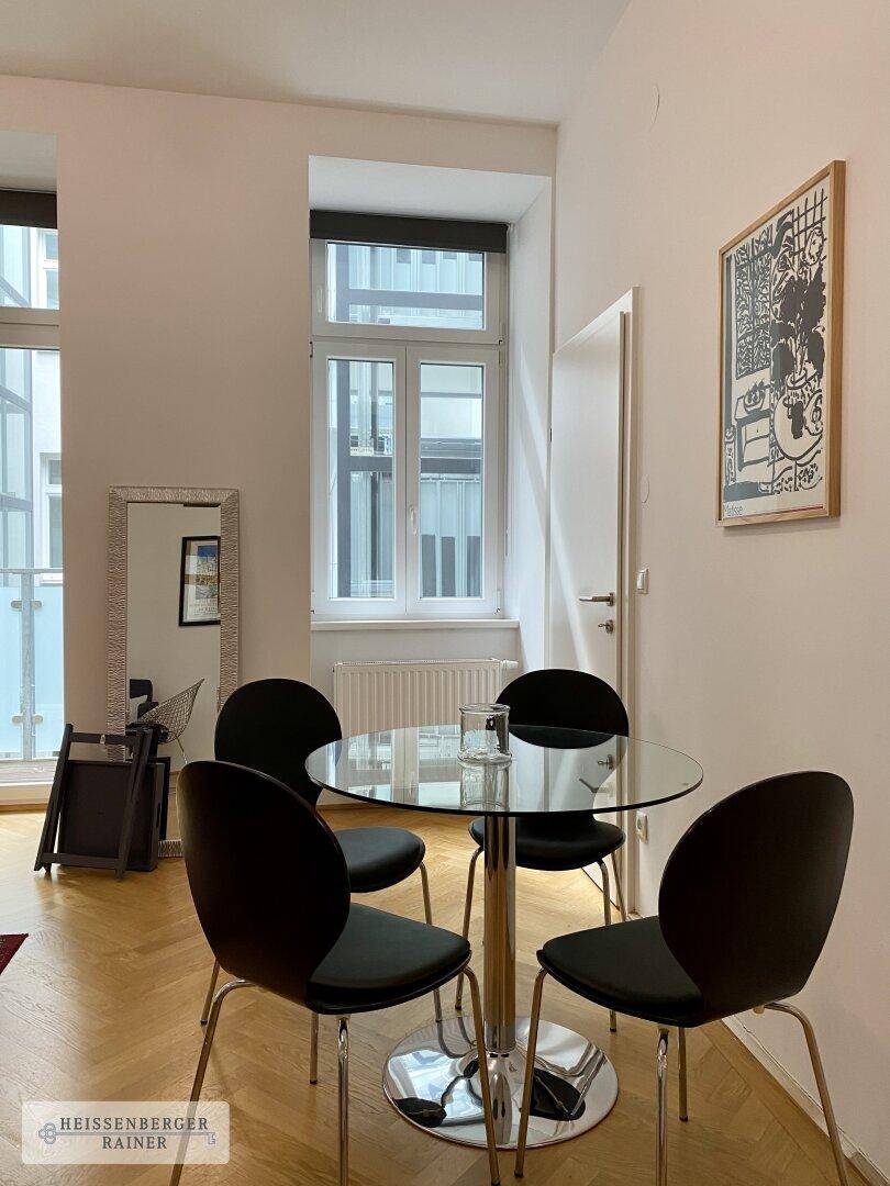 Essbereich mit kleinem Glastisch und Stühlen, hell durch große Fenster, ideal für Mahlzeiten.