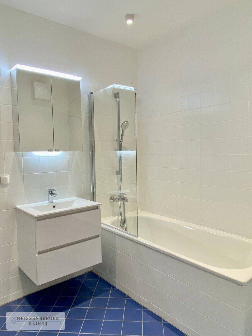 Modernes Badezimmer mit Badewanne, Waschtisch, Spiegelschrank und blauen Bodenfliesen.