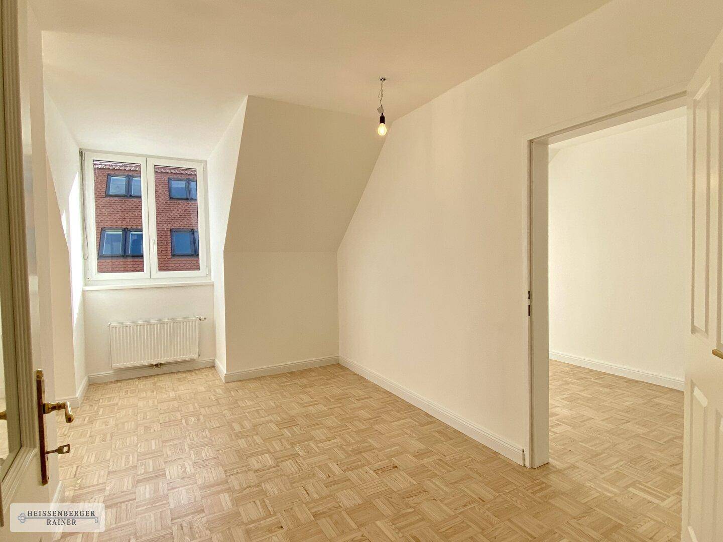 Helles Zimmer im Dachgeschoss mit Parkettboden, Schrägen und Blick auf gegenüberliegende Gebäude.