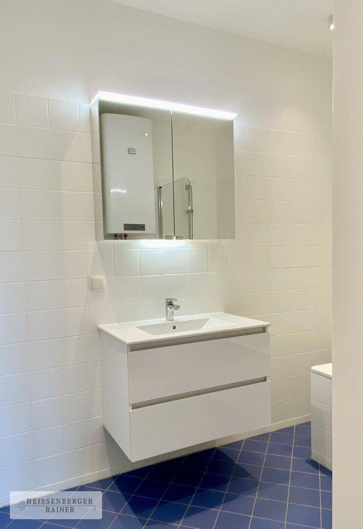 Helles Badezimmer mit schwebendem Waschtisch, beleuchtetem Spiegelschrank und blauen Bodenfliesen.