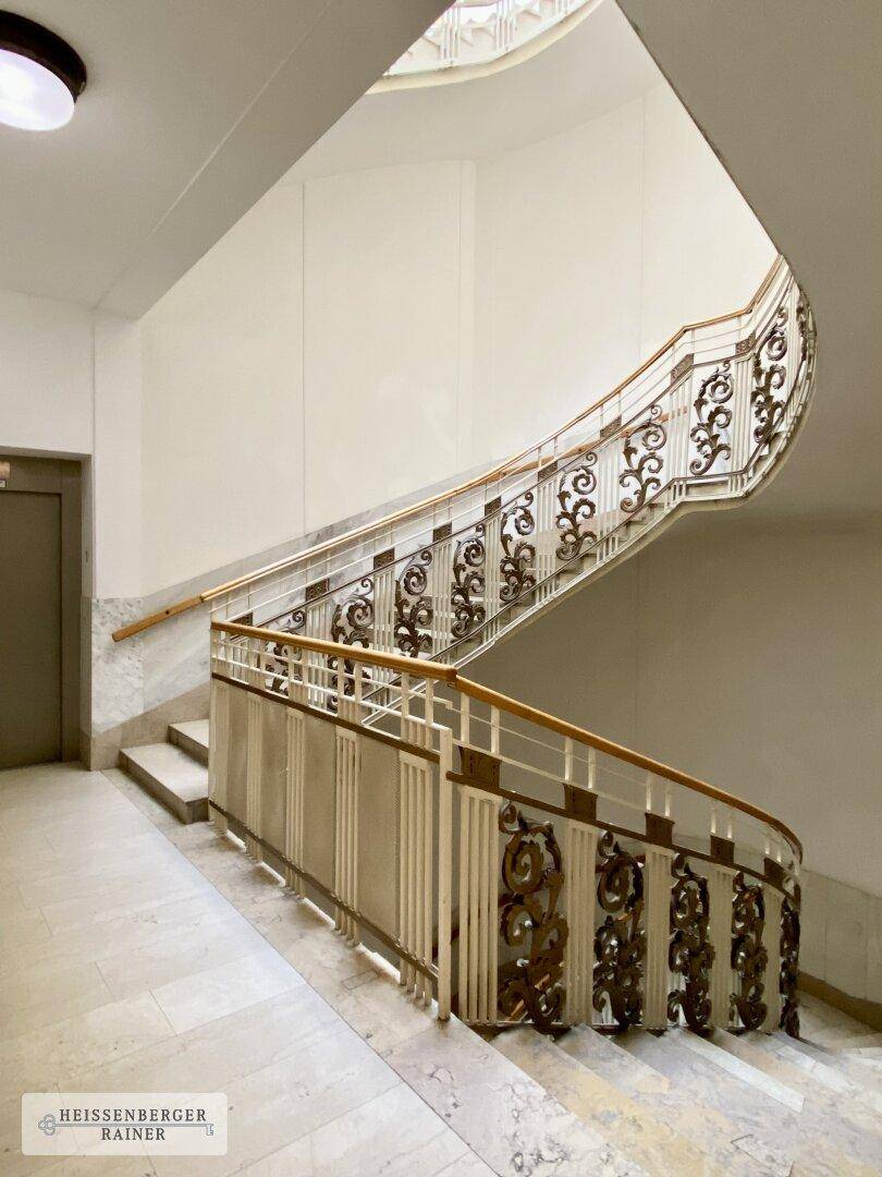 Elegantes Treppenhaus mit kunstvollen Geländern, Marmorstufen und einem Aufzug im Hintergrund.