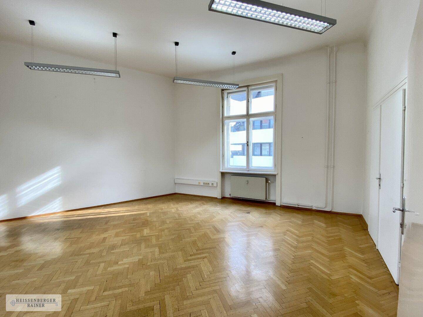 Heller Büroraum mit Fischgrätparkett, einem großen Fenster und moderner Deckenbeleuchtung.