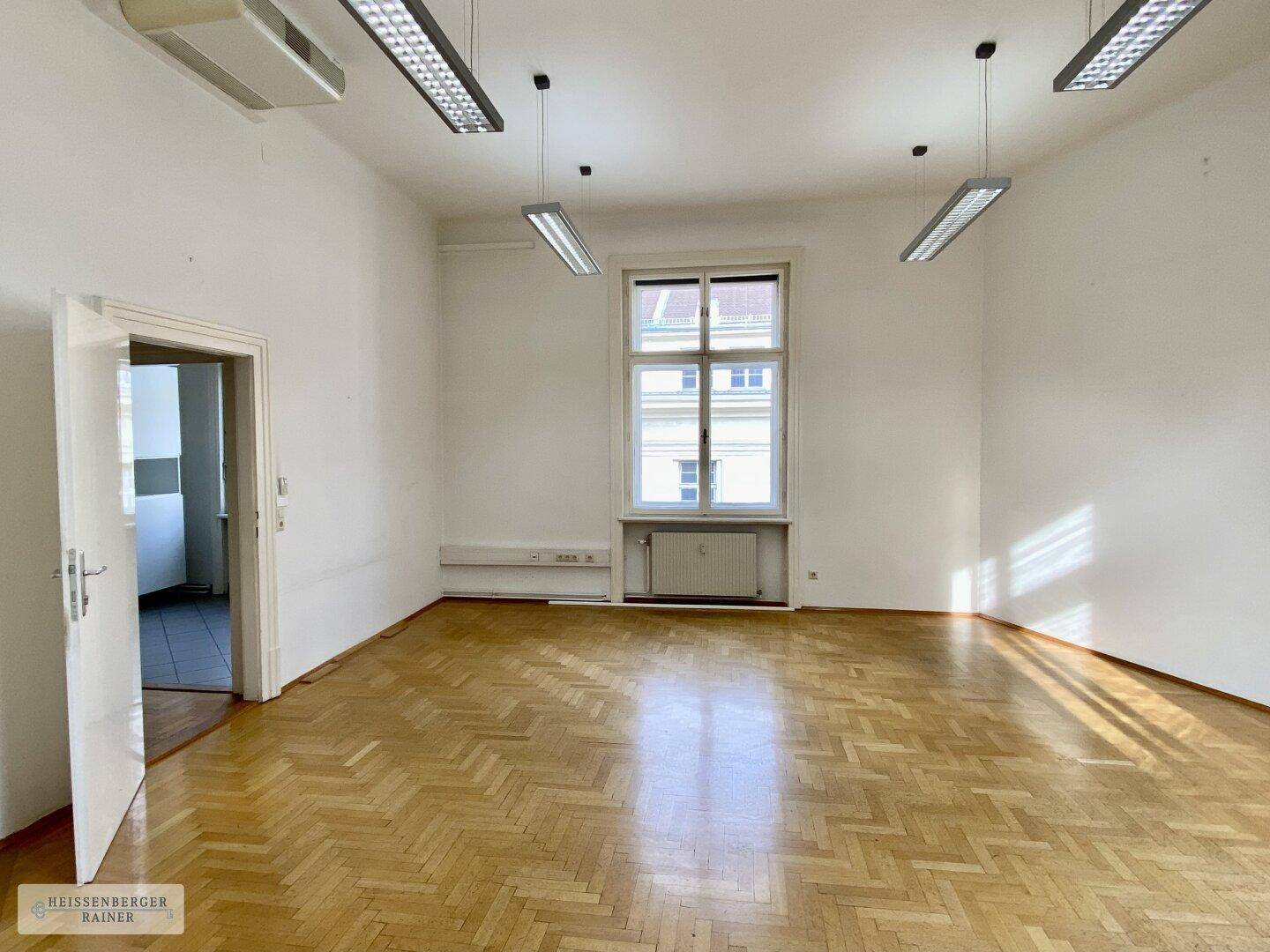 Leerer Büroraum mit Fischgrätparkett, einem Fenster und einem Durchgang zu einem weiteren Bereich.