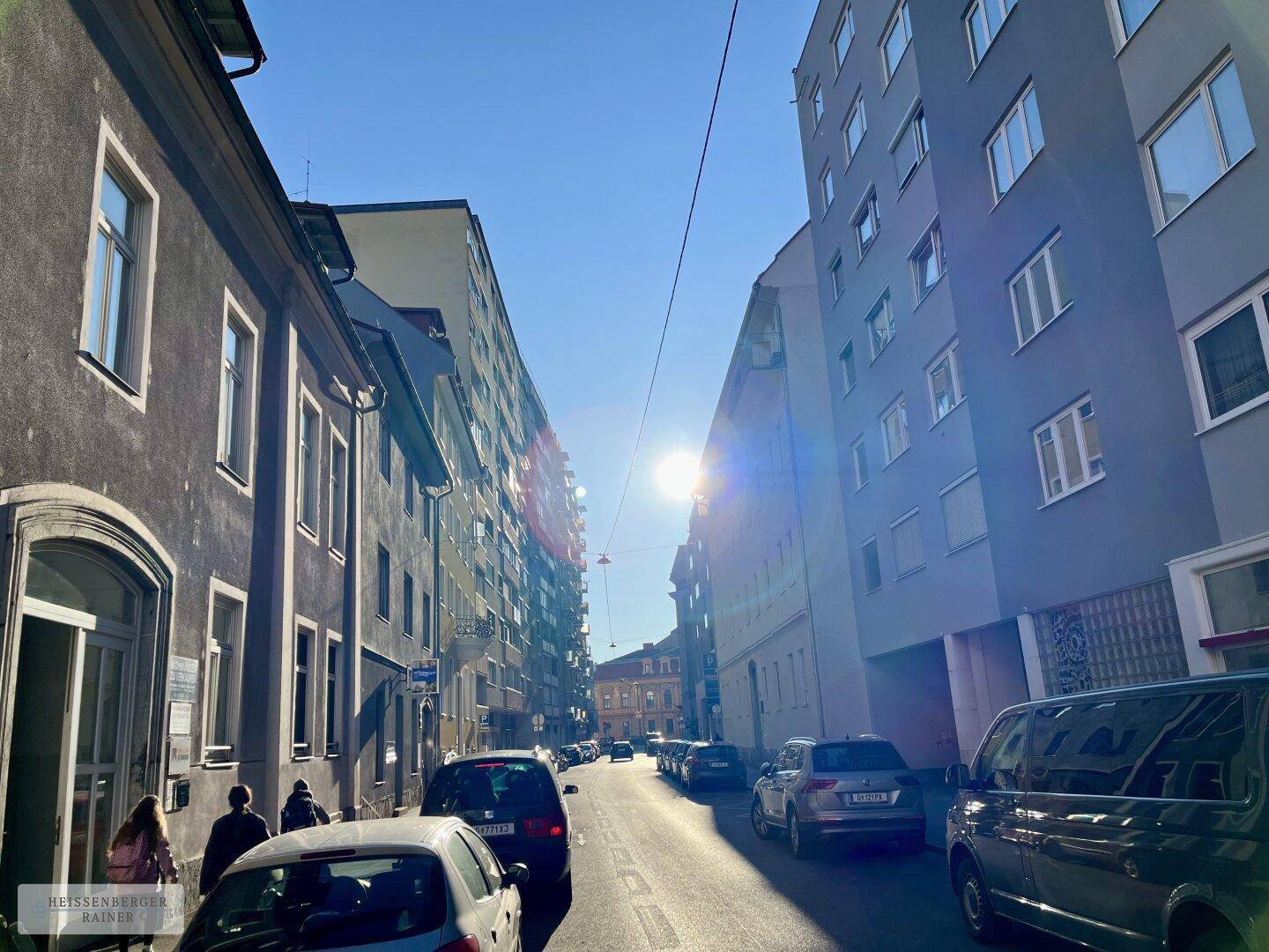 Sonnige Stadtstraße mit Gebäuden auf beiden Seiten und parkenden Autos unter blauem Himmel.