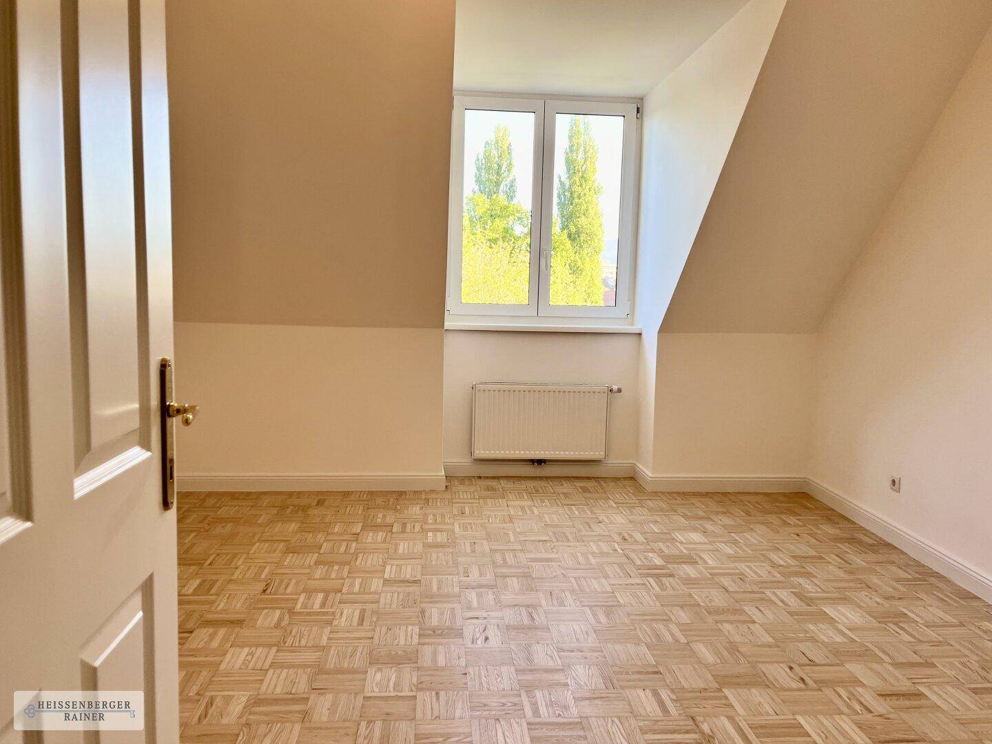 Leeres Zimmer mit hellem Parkettboden, Dachschrägen und einem Fenster mit Blick ins Grüne.
