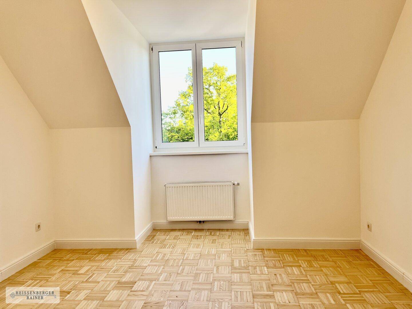 Helles Dachgeschosszimmer mit Parkettboden, schrägen Wänden und Blick ins Grüne.
