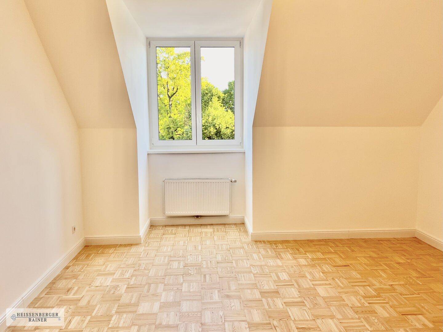 Geräumiges Dachgeschosszimmer mit Parkettboden, schrägen Wänden und Blick ins Grüne.