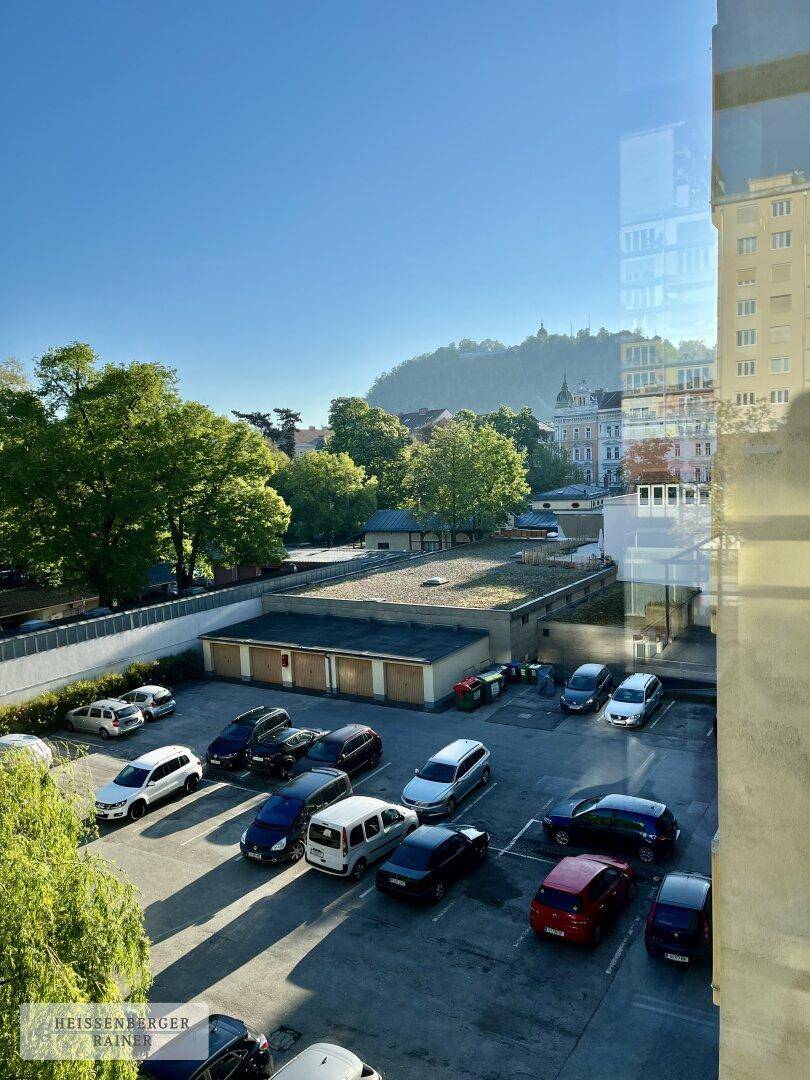 Blick auf einen Parkplatz mit umliegenden Gebäuden und einem bewaldeten Hügel im Hintergrund.