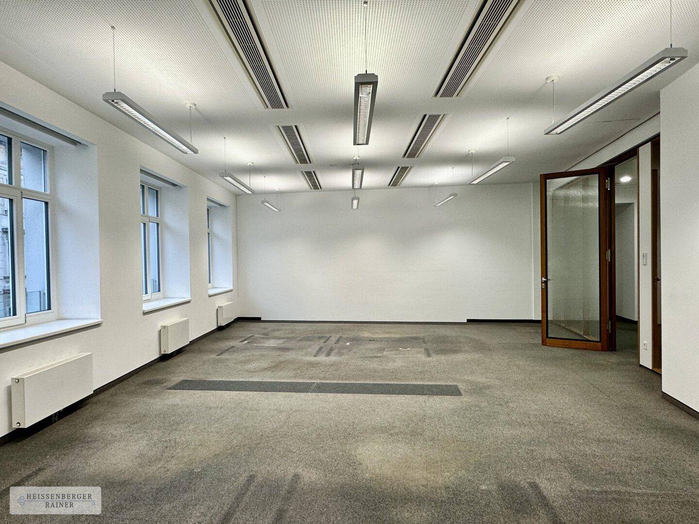 Helles Büro mit großen Fenstern, Teppichboden und abgehängter Decke mit integrierter Beleuchtung.