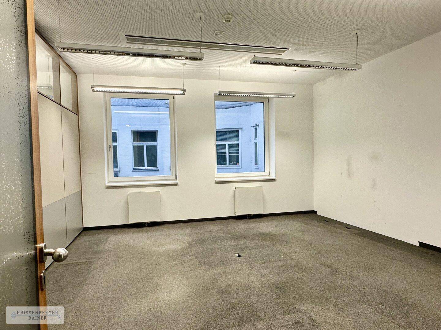 Kompaktes Büro mit Teppichboden, zwei Fenstern und integrierten Schränken.
