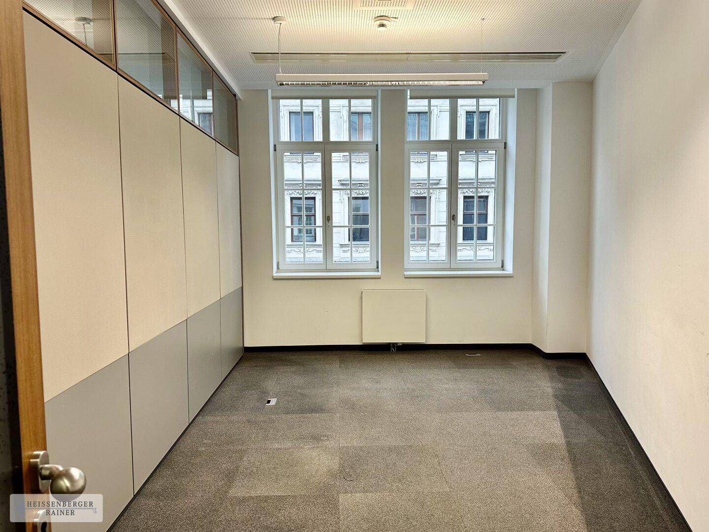 Büro mit Teppichboden, hohen Fenstern und einer Wand aus Einbauschränken.