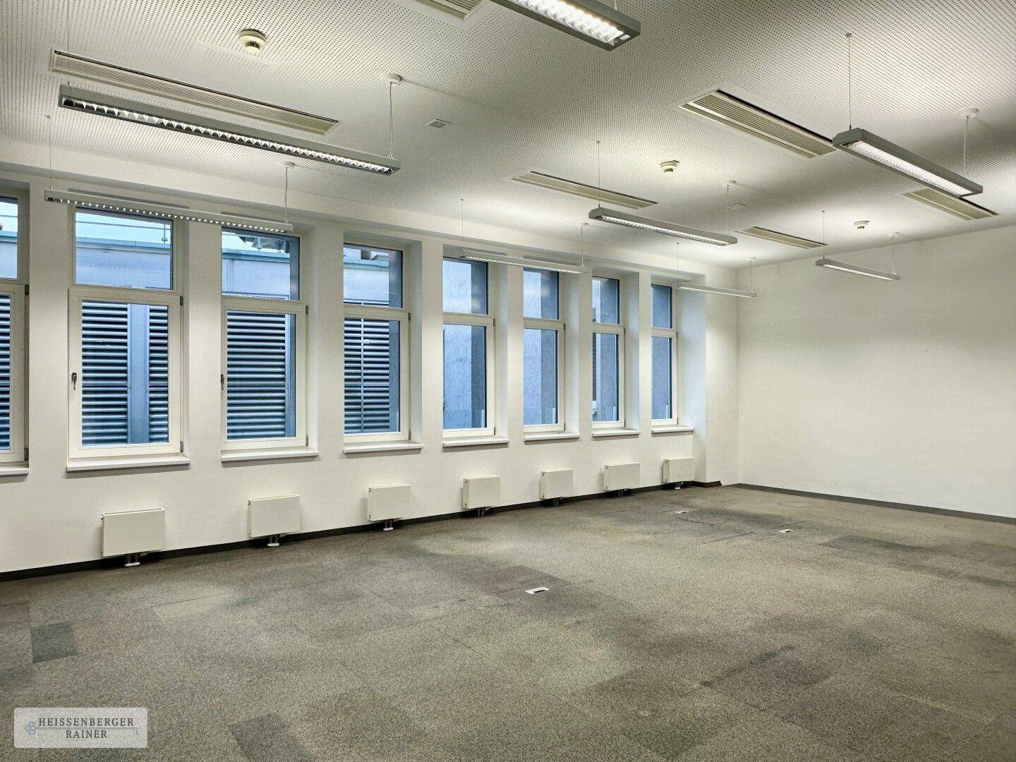 Geräumiges Büro mit vielen Fenstern, Teppichboden und effizienter Deckenbeleuchtung.
