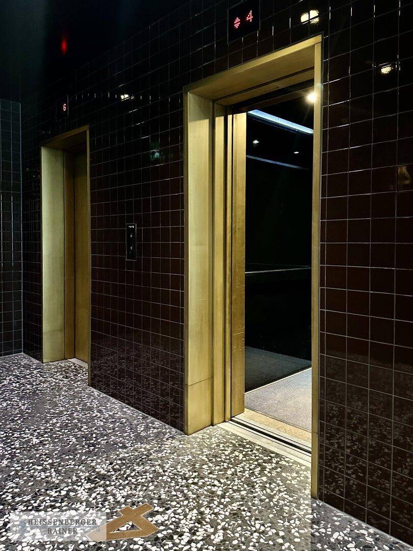 Aufzugsbereich mit dunklen Fliesenwänden, goldenen Aufzugstüren und Terrazzoboden.