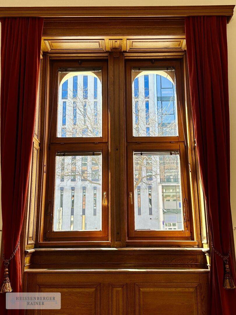 Detailansicht eines großen Holzfensters mit roten Vorhängen, das einen Blick auf moderne Architektur bietet.