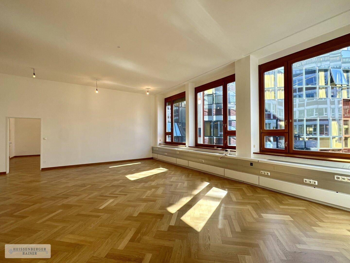 Geräumiger, leerer Büroraum mit Fischgrätparkett und großen Fenstern, die viel Tageslicht hereinlassen.