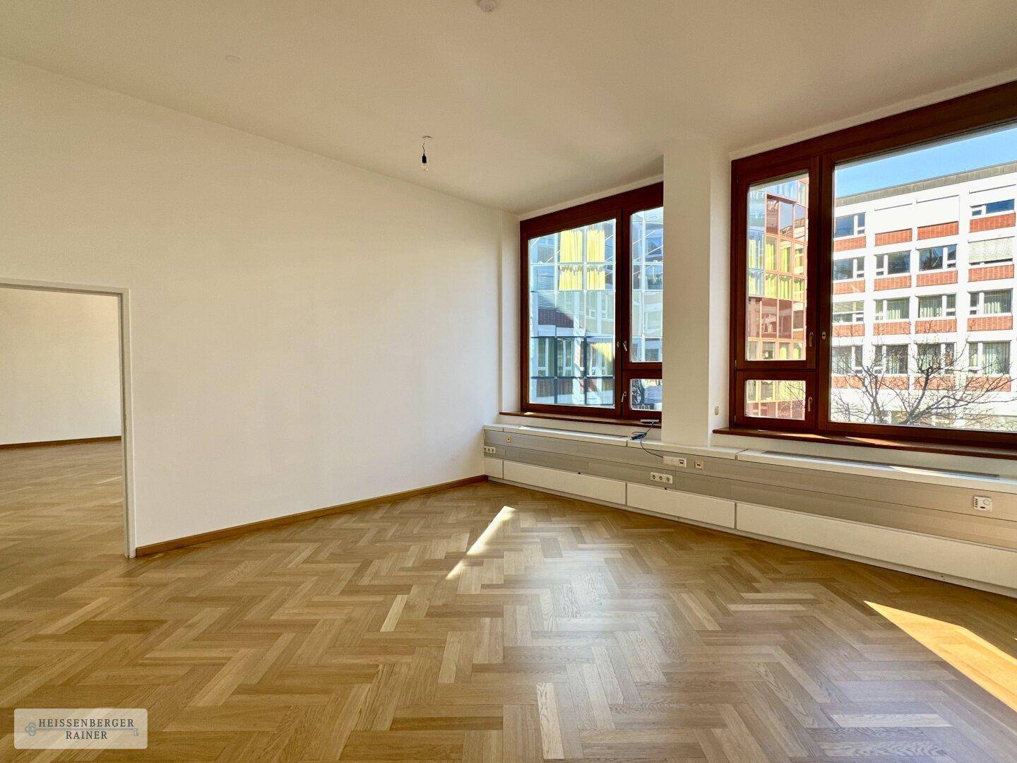 Leerer Büroraum mit hellem Fischgrätparkett und großen Fenstern, die viel Sonnenlicht hereinlassen.