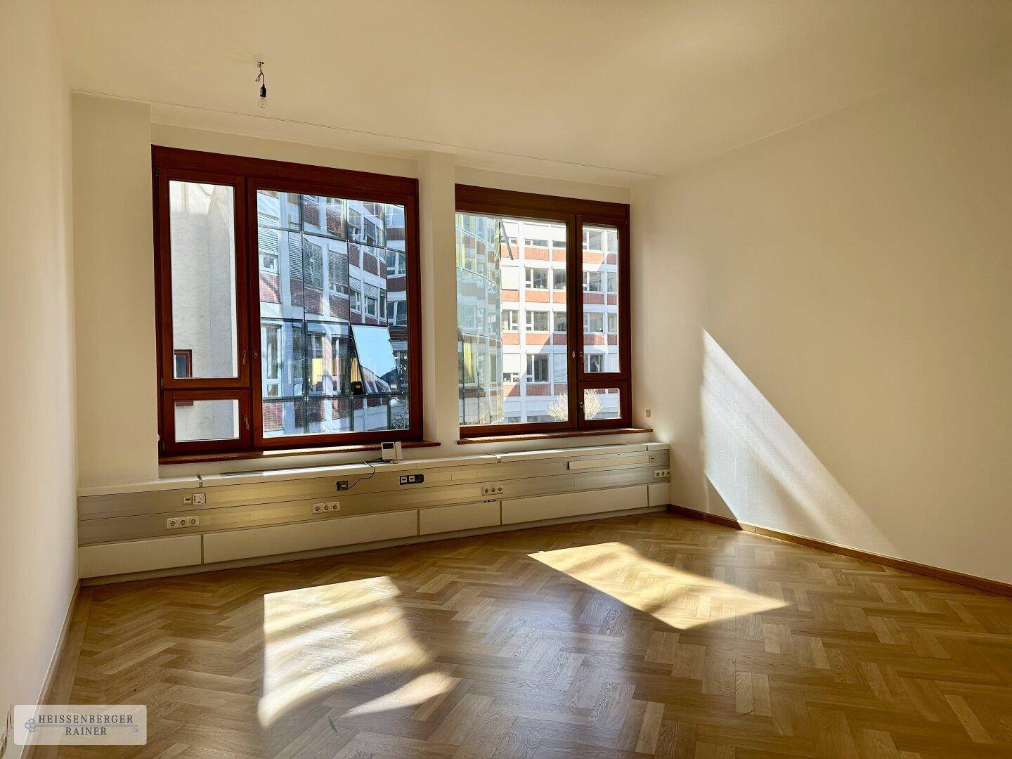 Heller Büroraum mit Fischgrätparkett und zwei großen Fenstern, die einen Blick auf die Stadt ermöglichen.