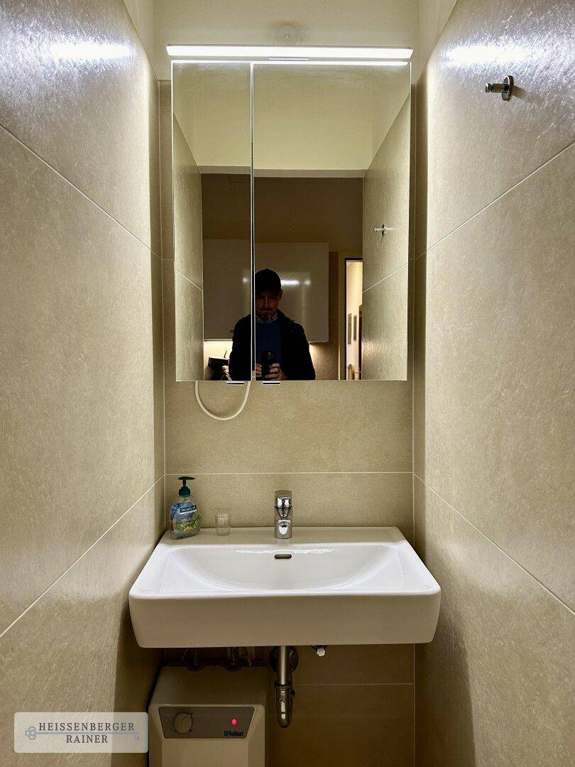 Kleines Badezimmer mit Waschbecken, Spiegel und beigefarbenen Fliesen.