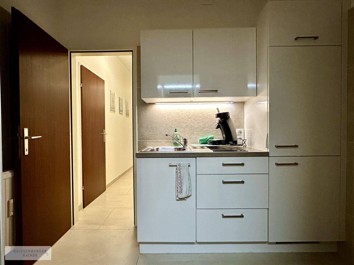 Kleine, moderne Küchenzeile mit weißen Schränken, Spüle und Kaffeemaschine.