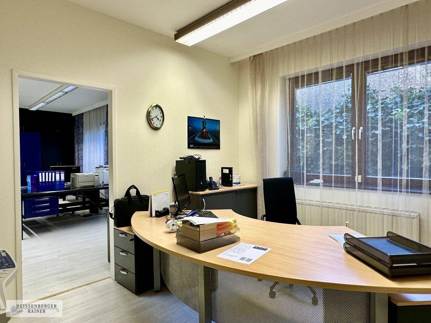 Einzelbüro mit Holzschreibtisch, Fenster und Blick in einen weiteren Büroraum.