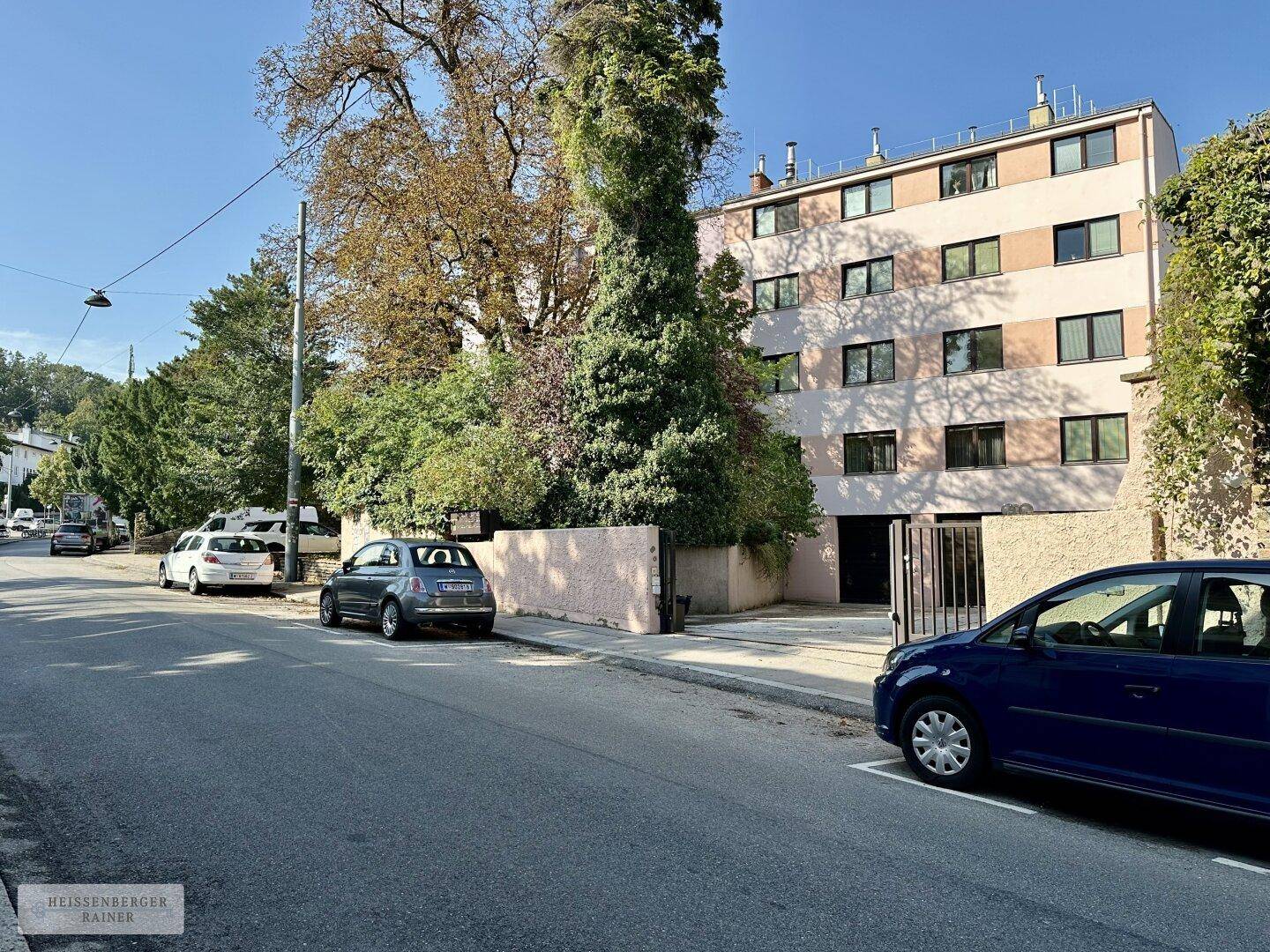 Mehrstöckiges Gebäude an einer Straße mit parkenden Autos und Bäumen.