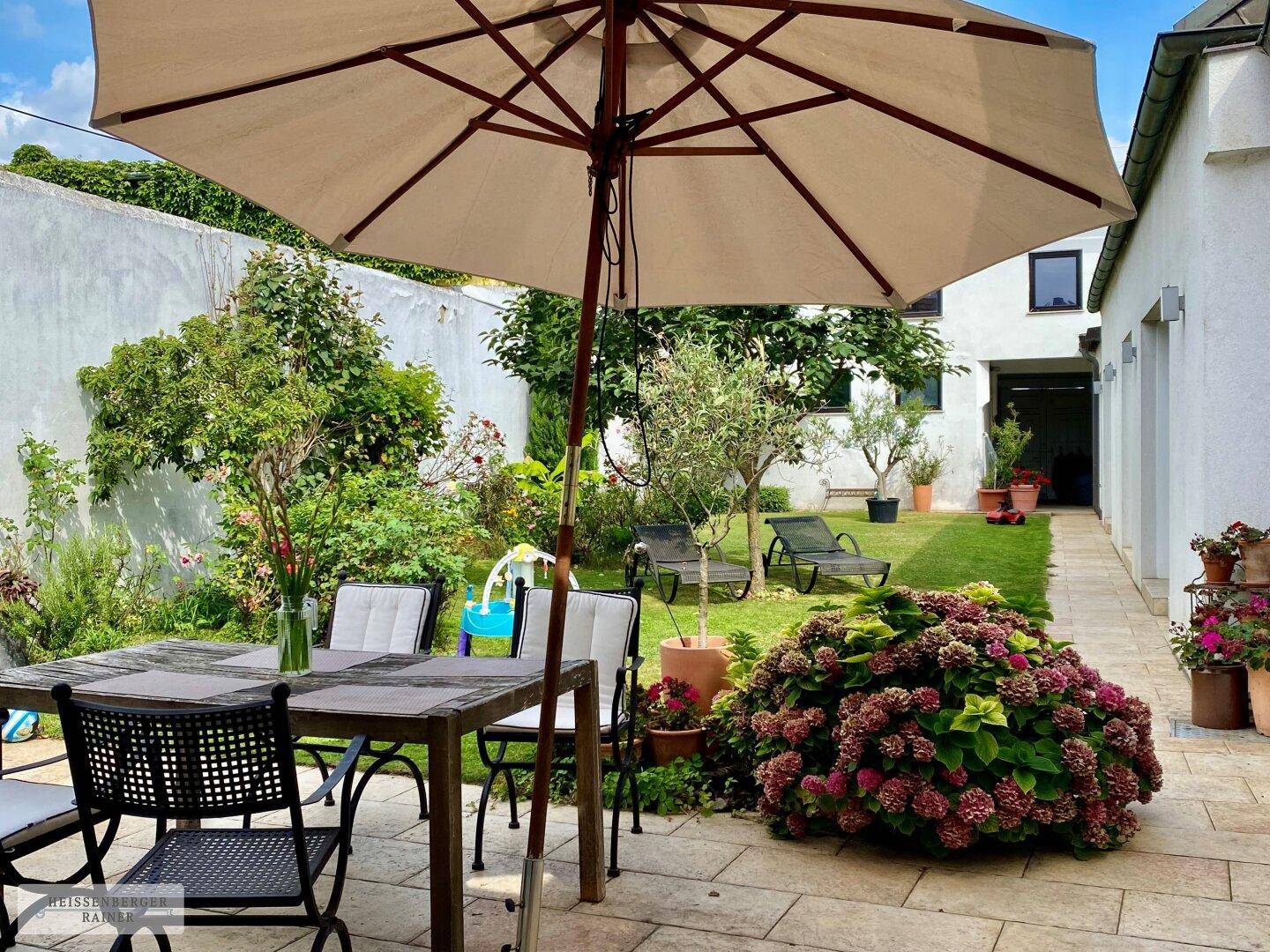 Gemütliche Terrasse mit Esstisch und Sonnenschirm, umgeben von üppigem Grün und Pflanzen.