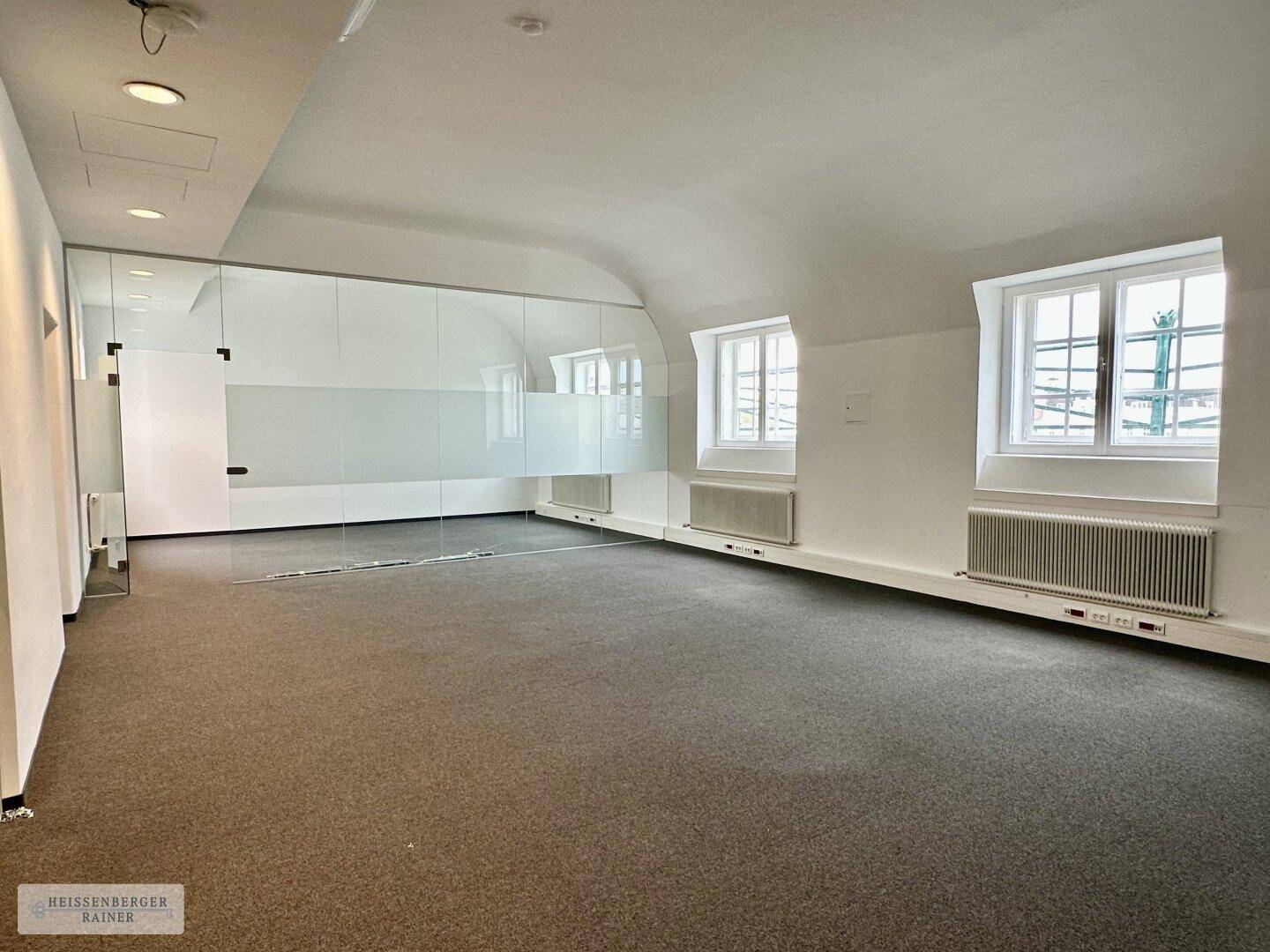 Geräumiges Büro mit Teppichboden, weißen Wänden und einer modernen Glasabtrennung, beleuchtet durch Bogenfenster.