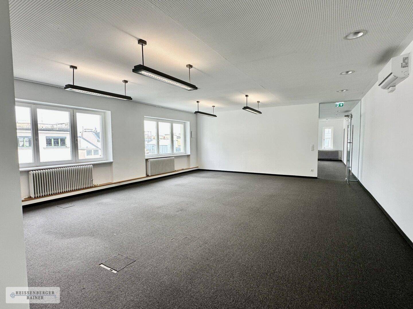 Großer offener Bürobereich mit Teppichboden, vielen Fenstern und moderner Deckenbeleuchtung für eine helle Arbeitsumgebung.