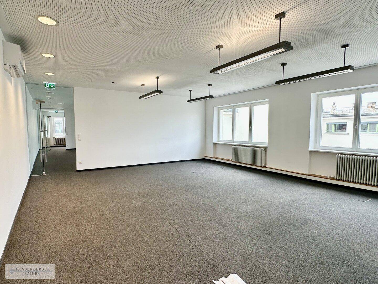 Weitläufiger Büroraum mit Teppichboden, zahlreichen Fenstern und effizienter Deckenbeleuchtung, ideal für Großraumbüros.