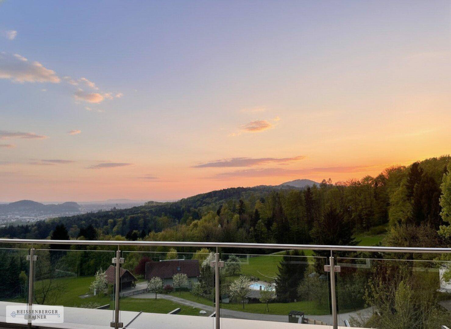 Malerischer Sonnenuntergang über der grünen Landschaft, gesehen von einer modernen Terrasse mit Glasgeländer.