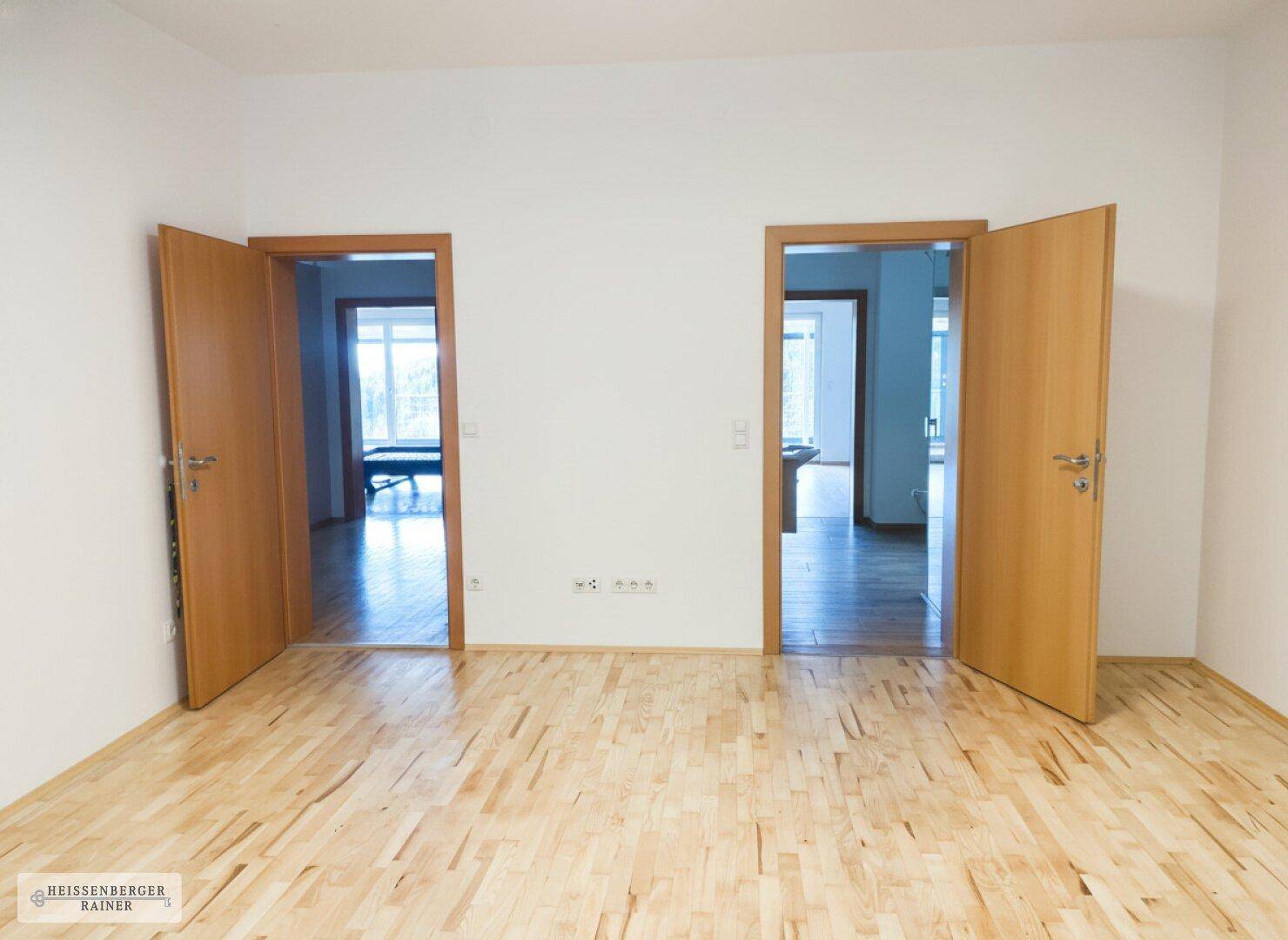 Heller Raum mit Holzboden und zwei offenen Türen, die zu weiteren Zimmern führen.