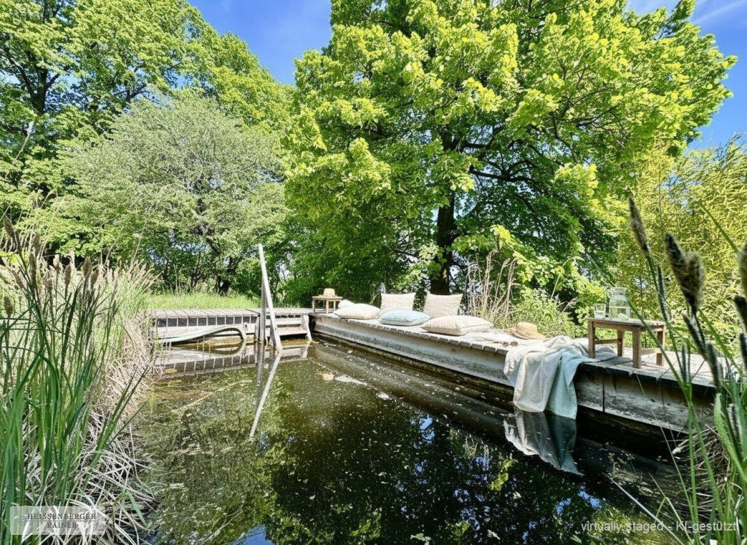 Natürlicher Teich mit Holzdeck und Sitzgelegenheiten, umgeben von üppiger Vegetation.