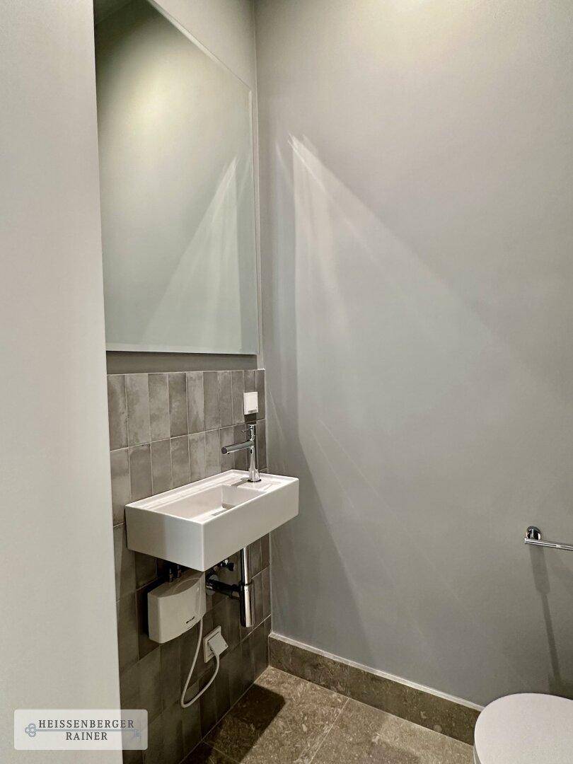Modernes Gäste-WC mit kleinem Waschbecken, Spiegel und grauen Fliesen an der Wand.