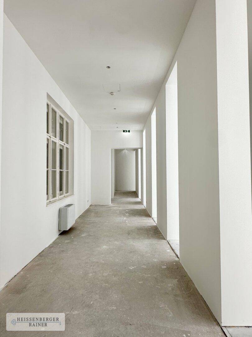 Langer, heller Flur mit weißen Wänden und Rohbetonboden, der zu weiteren Räumen führt.