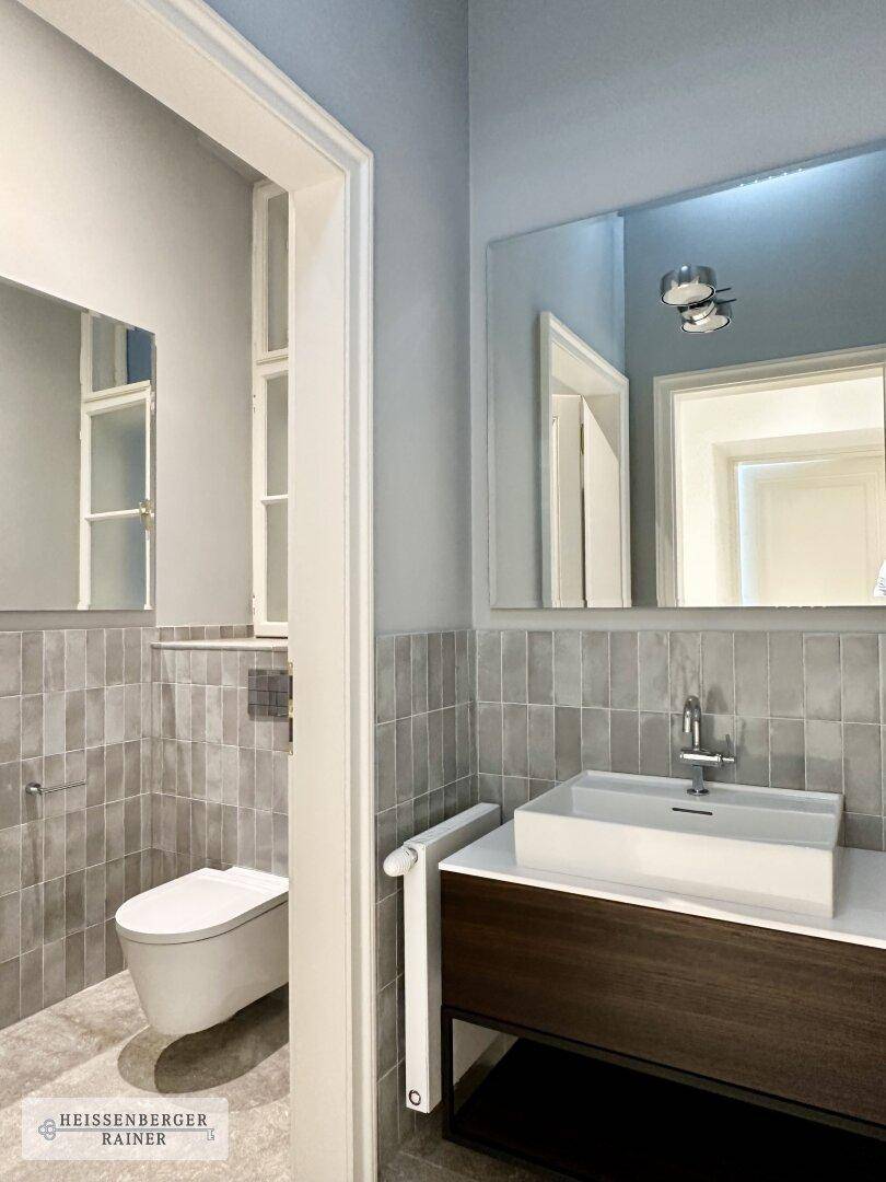 Modernes Badezimmer mit grauen Fliesen, wandhängendem WC und stilvollem Waschtisch mit Spiegel.