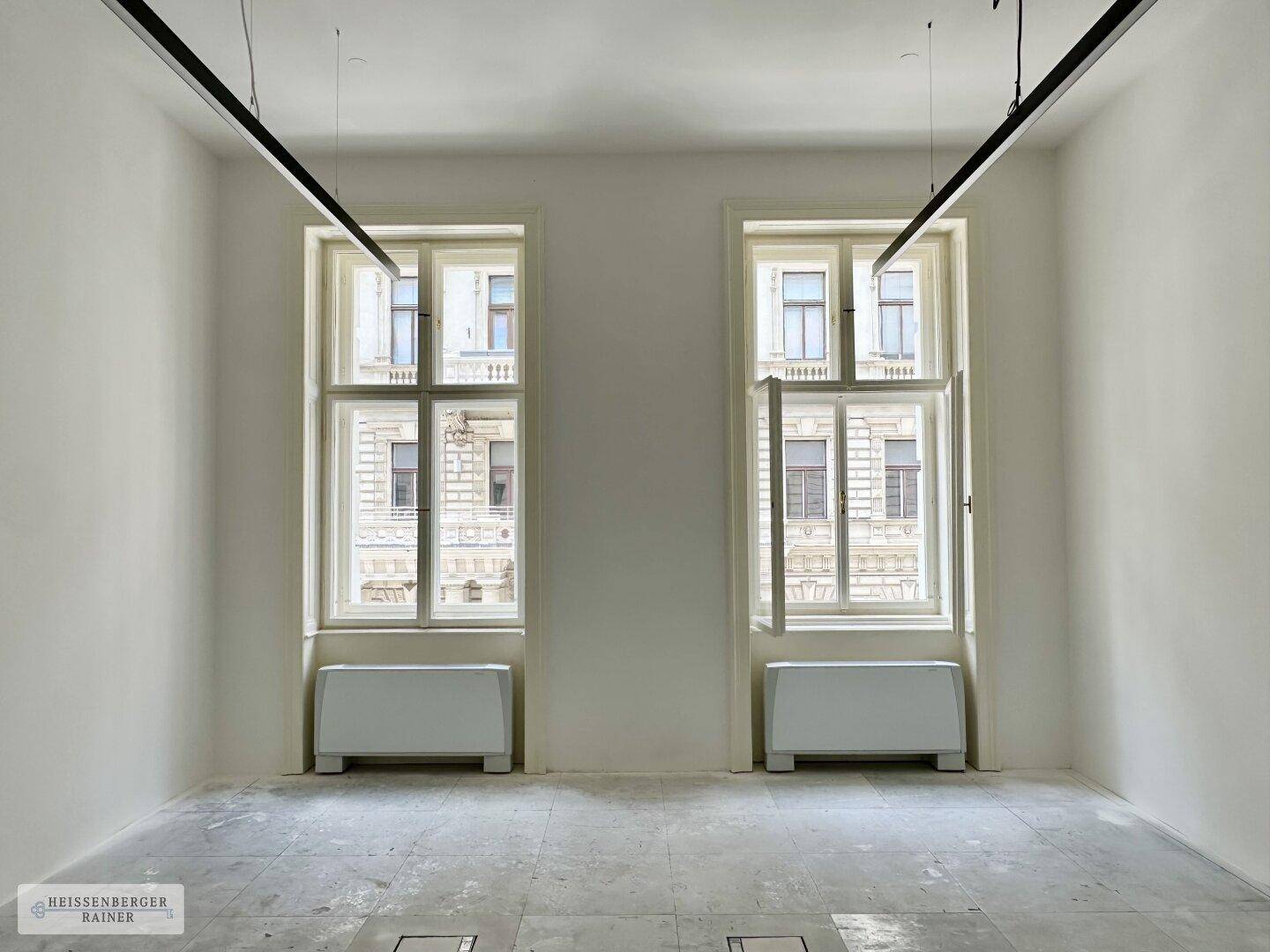 Ein heller, leerer Raum mit zwei hohen Fenstern und modernen Deckenleuchten.