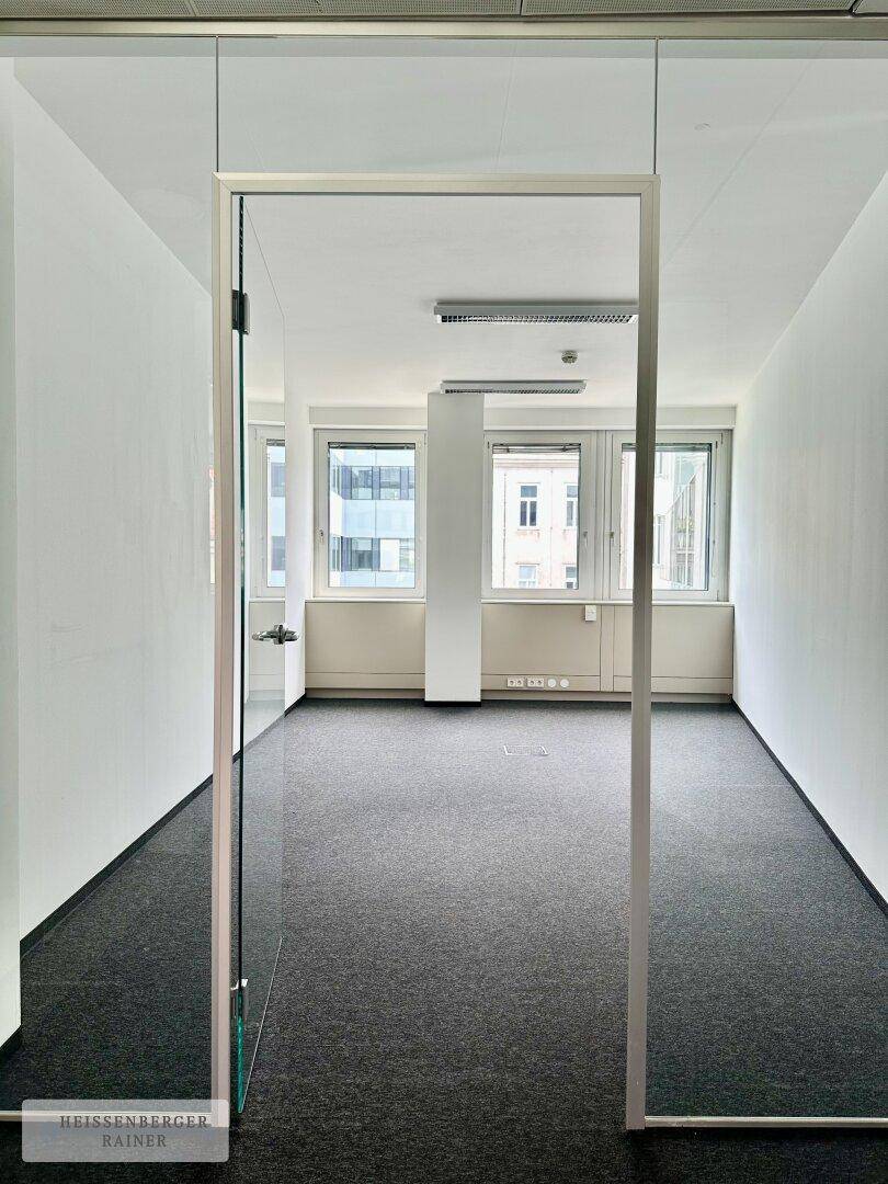 Blick durch eine Glastür in einen hellen Büroraum mit Teppichboden und Fenstern, die Tageslicht spenden.
