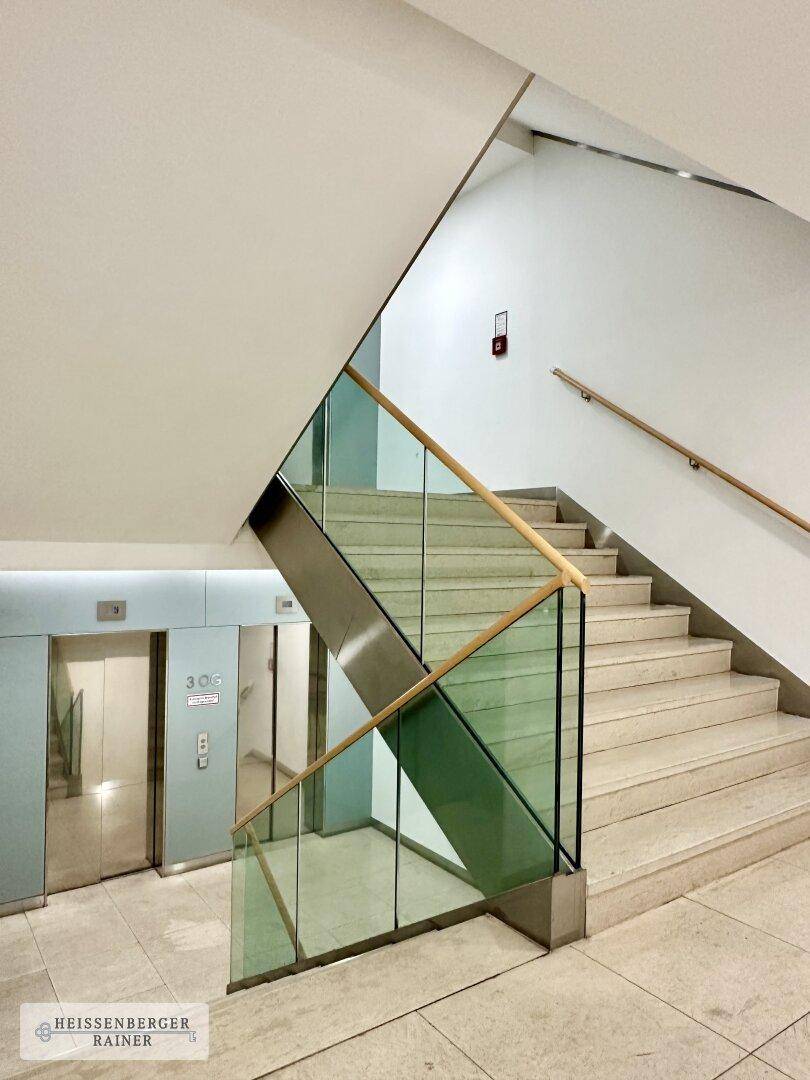 Modernes Treppenhaus mit Glasgeländer und Holzhandlauf, direkt neben den Aufzügen gelegen.
