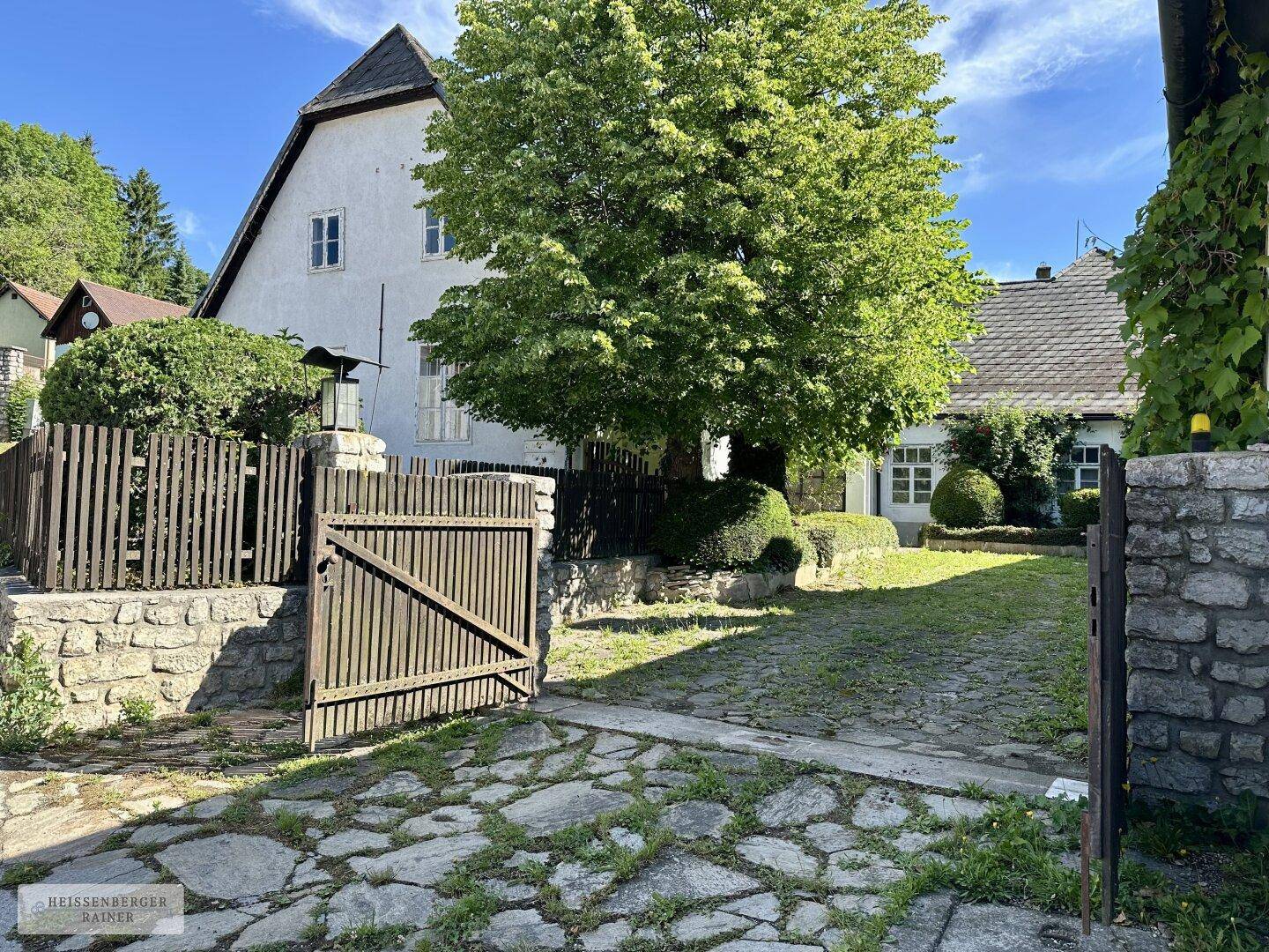 Einfahrt zum Anwesen mit Holztor, Steinmauer und einem großen Baum im Vorgarten.